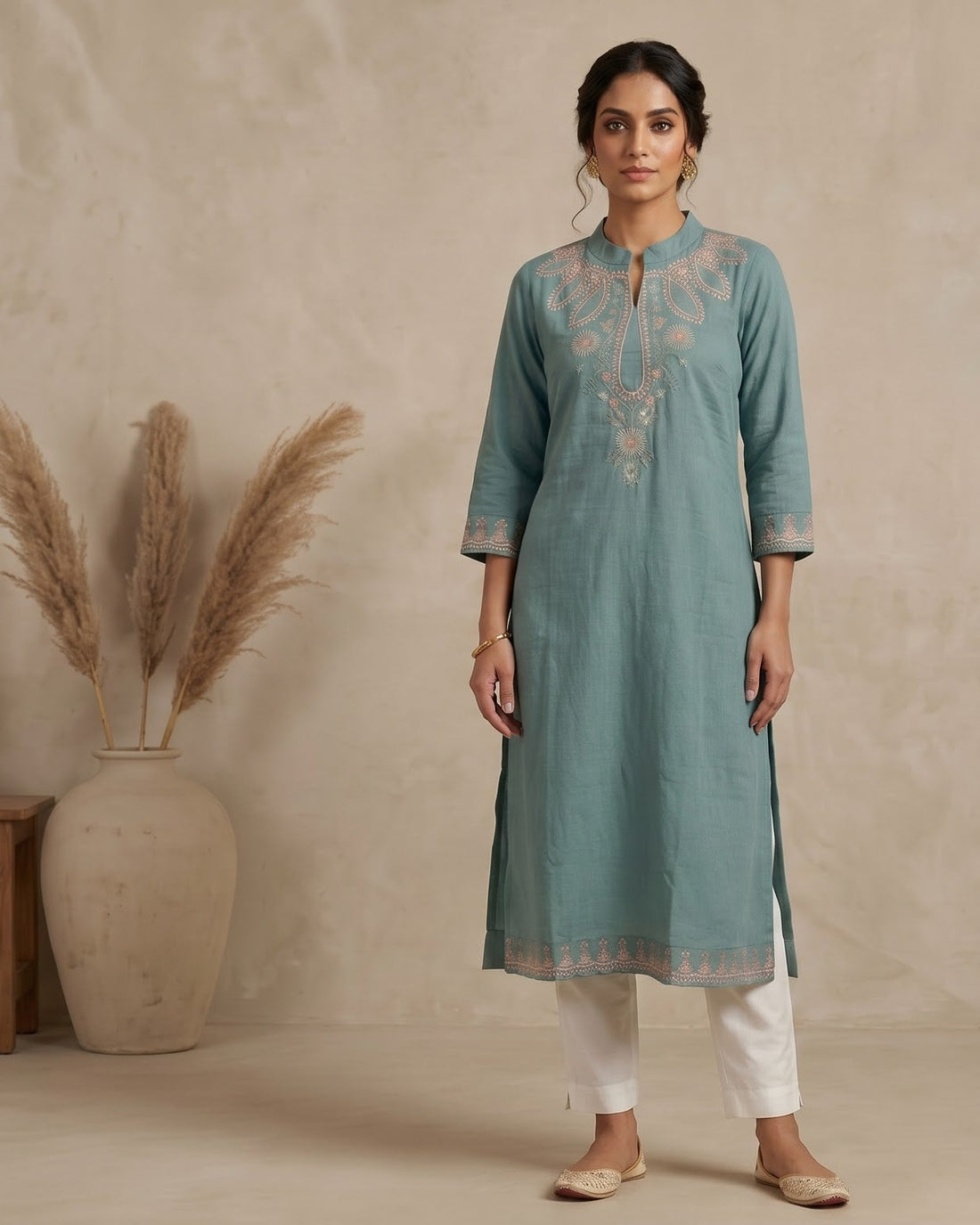 A woman stands in a studio wearing a light teal-blue kurta with beige embroidery, paired with white pants and beige shoes. The background is a textured beige wall with a large vase of dried grass on the left and a small wooden bench on the right.