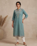 A woman stands in a studio wearing a light teal-blue kurta with beige embroidery, paired with white pants and beige shoes. The background is a textured beige wall with a large vase of dried grass on the left and a small wooden bench on the right.