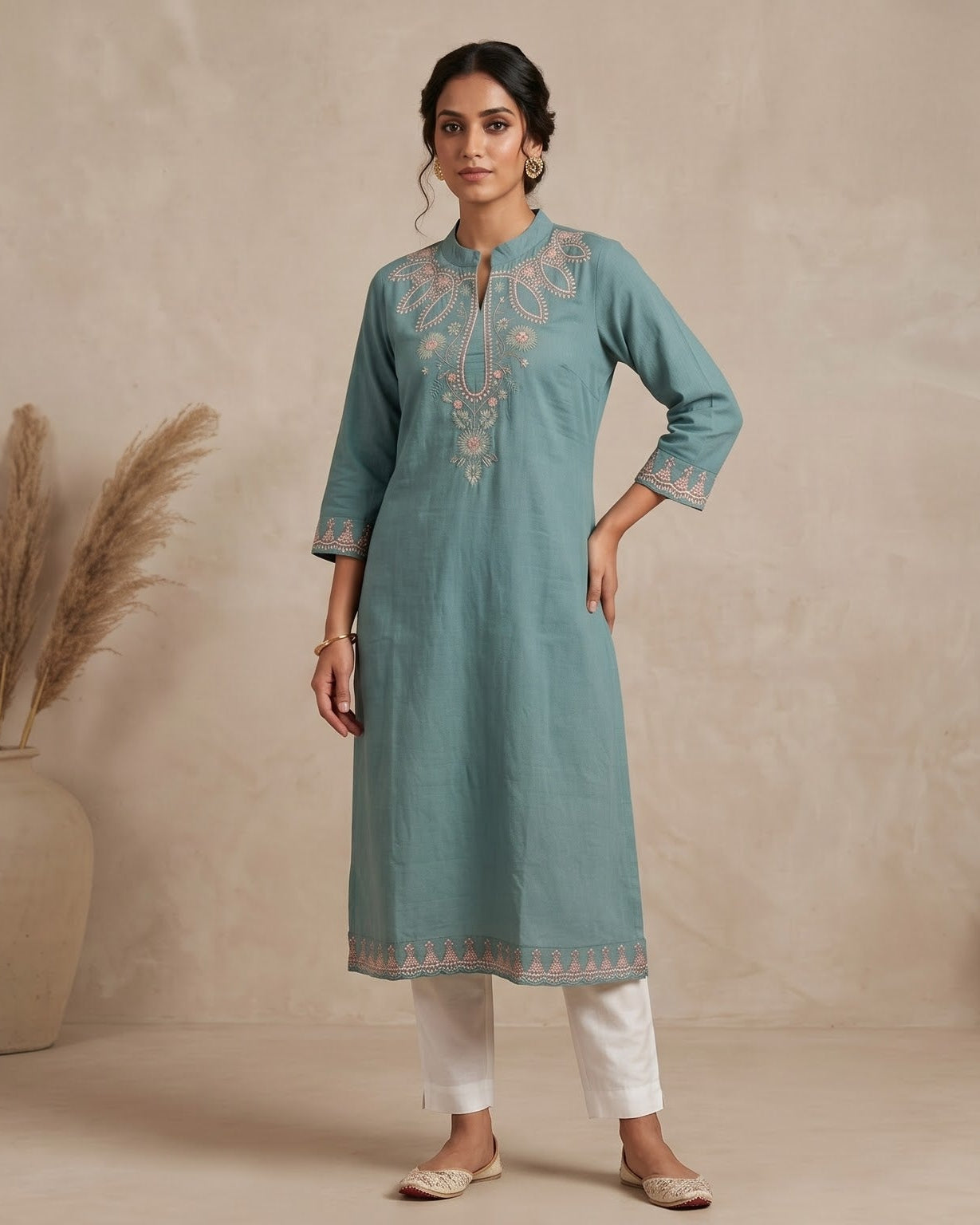 A woman stands in a studio wearing a light teal-blue kurta with beige embroidery, paired with white pants and beige shoes. The background is a textured beige wall with a large vase of dried grass on the left and a small wooden bench on the right.