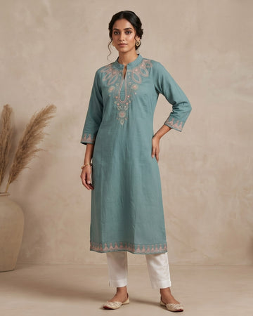 A woman stands in a studio wearing a light teal-blue kurta with beige embroidery, paired with white pants and beige shoes. The background is a textured beige wall with a large vase of dried grass on the left and a small wooden bench on the right.